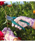 Hands wearing floral gloves using garden scissors, surrounded by plants with red and orange flowers in the background.