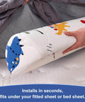 Toddler bed bumper with dinosaur print being placed under gray bed sheet; 2 Pack Premium Foam Toddler Bed Bumpers With Non Slip Rails For Twin Queen And King Beds Cozy Odor Free Comfort Machine Washable Covers Easy Care 52 By 7 By 5 Inches Safe Sleep Aid For Peaceful Nights.