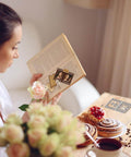 Woman reading a book with Awesome Versatile Die Cut Set For Card Making And Scrapbooking; floral decor and pastries on table.