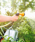 Person using a garden hose with brass quick connect kit spraying water on plants with a wheelbarrow in the background.