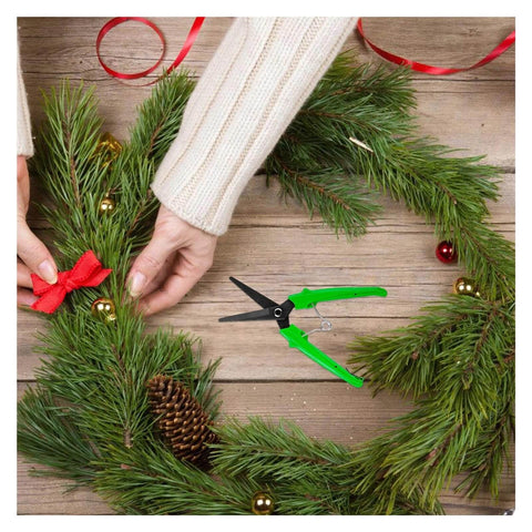 Person crafting holiday wreath with green-handled garden scissors, pine needles, pinecone, and red ribbon on wooden table.