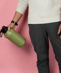 Person holding a green water bottle against a pink background, wearing a white sweatshirt and dark pants.