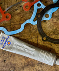 Grey Cut Gasket Coating Sealant For Tough Torque Engines, Vibration Resistant Silicone Sealant For Paper Cork And Rubber Gaskets, Extends Gasket Life And Prevents Blowouts, Fills Scars, Tack Free In 1 Hour, 2 Oz tube on wooden background, with various colored gaskets beside it.