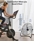 Cyclist using exercise bike near 20 Inch Heavy Duty Floor And Wall Mount Fan For Industrial Cooling With 5750 CFM High Velocity Three Speed And 360 Degree Tilt Head Black UL Listed For Garage Workshop Office And Home.