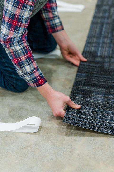 Person installing carpet tile with Heavy Duty Double Sided Tape For Carpet Tiles And Vinyl Flooring, Ideal For Artificial Grass, Foam Gym Tiles And Rubber Interlocking Gym Tiles, Easy Installation With Ultra Strong Grip And Industrial Strength Bond No Mess