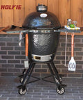 Black ceramic grill with side shelves on stand with wheels, set against a brick wall.