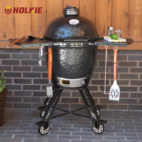 Black ceramic grill with side shelves on stand with wheels, set against a brick wall.