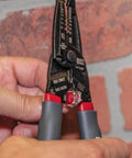 Close-up of hands using Kurve Wire Stripper Crimper Cutter Multi Tool for 10-26 AWG solid and 12-28 AWG stranded wire against a brick wall