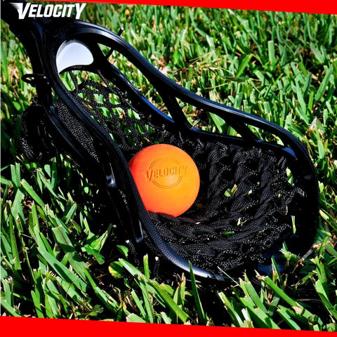 Close-up of a lacrosse stick head with an orange ball on grass, highlighting Velocity branding.