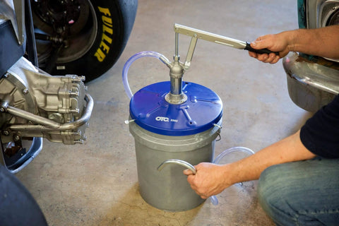 Person using OTC 2352 Lever Action Bucket Pump on 5-gallon pail with handle and hose visible in a garage setting.