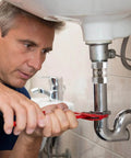 Person using wrench on plumbing beneath a sink with metal piping components.