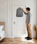 Person hanging a gray backpack on a white door in a modern bedroom with wooden floor and a beige bed.