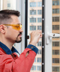 Person installing or adjusting a window handle with tools, wearing safety glasses and red protective clothing, buildings in background