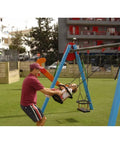 Adult pushing a child on a blue and red swing set in a playground with grass and buildings in the background.
