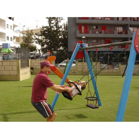 Adult pushing a child on a blue and red swing set in a playground with grass and buildings in the background.