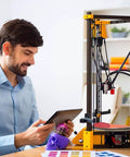 Man operating a 3D printer with a tablet, surrounded by printed objects and color samples on a desk in an office setting.