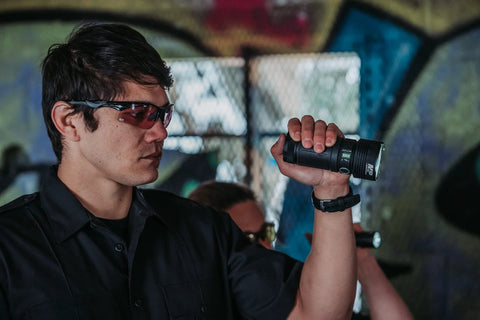 Man wearing Harrier Interchangeable Shooting Glasses with tinted lenses holding a flashlight in urban environment.