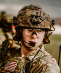 Soldier in camouflage helmet with matte black frames shooting glasses, ANSI Z87 safety and changeable lenses in outdoor setting.
