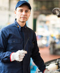 Mechanic in blue uniform and cap holding a dipstick next to an open car hood in a garage setting.