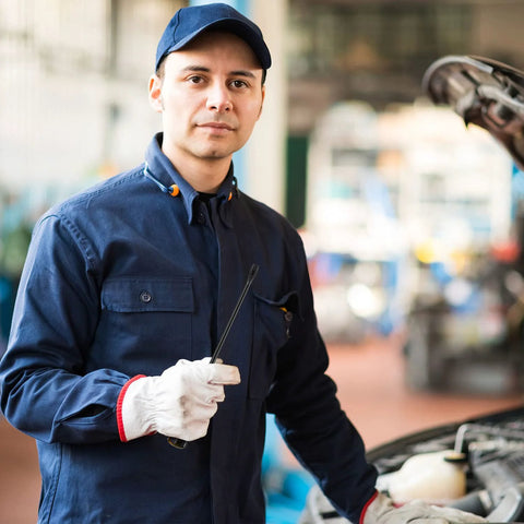 Mechanic in blue uniform and cap holding a dipstick next to an open car hood in a garage setting.