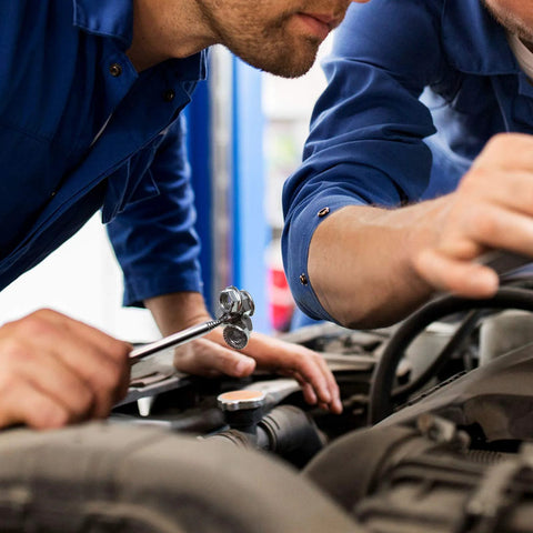 Mechanics using tools under the hood of a car in an auto shop, wearing blue uniforms, focused on engine repair work.