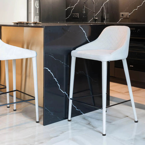 Two modern white bar stools with metal frames in a kitchen setting, contrasting against a black marble island counter.