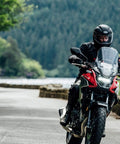 Motorcyclist riding through scenic forest road with Essentials Micro XSystem 3.0 Plus Electronic Automatic Motorcycle Chain Lubrication System visible.