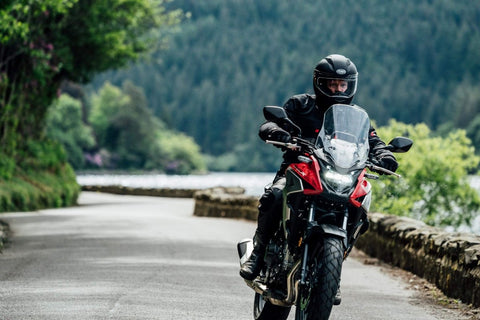 Motorcyclist riding through scenic forest road with Essentials Micro XSystem 3.0 Plus Electronic Automatic Motorcycle Chain Lubrication System visible.