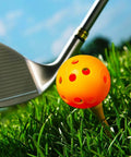 Orange Practice Golf Training Ball on tee, close-up view, with a golf club on grass. Limited Flight 40mm Hollow Plastic Colored Airflow Golf Balls.