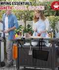 Two people using a grill station with various camping essentials, featuring magnetic grill hooks holding tools.
