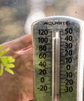 Close-up of an outdoor thermometer displaying temperatures in Fahrenheit and Celsius next to a green plant leaf.