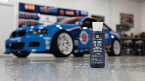 Permatex 81950 Ultra Slick Engine Assembly Lube, 4 oz. , Red bottle in front of a blue racing car in a garage setting.