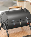 Black portable charcoal grill with wooden handle on a wooden table, features two latches and chimney, designed for outdoor cooking.