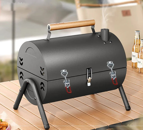 Black portable charcoal grill with wooden handle on a wooden table, features two latches and chimney, designed for outdoor cooking.