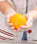 Person wearing Powder Free Vinyl Gloves 4.3 Mil Clear Latex Free For Food Service Cleaning, washing an orange under tap water.