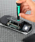 Using the 104 Piece Home Repair Tool Set With Easy Carrying Pouch For DIY Projects And Everyday Fixes Includes Hammer Pliers Magnetic Screwdrivers And Bits Wrenches Utility Knife Level And More In A Turquoise Portable Kit, a hand uses a screwdriver to open a portable gaming device on a cutting mat.