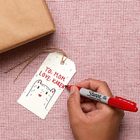 Hand using fine point permanent marker to write on a gift tag, showing bold, precise lines with quick-drying, odorless ink.