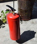 A red metal fire extinguisher with a black handle, placed on a concrete surface near decorative planters.