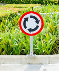 Roundabout sign on metal pole in front of green hedge and plants