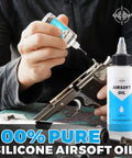 Person applying Airsoft Oil 4 oz & 1 oz Needle Oiler 100% Silicone Air Gun Oil on airsoft gun, showing both bottle sizes.