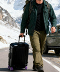 Man walking with suitcase using Silicone Luggage Wheel Covers Protector Set For Travel And Office Chair Casters Wear Resistant Anti Noise Shock Absorption Soft Stretchable Covers That Reduce Drag And Vibration Quiet Purple on a snowy mountain road