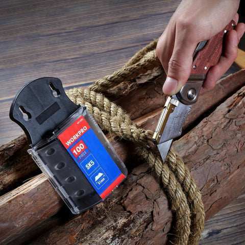 Close-up of SK5 Steel Utility Knife Blades 100 Pack with Dispenser next to utility knife cutting thick rope on wood surface