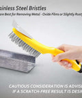 Hand holding stainless steel wire brush with metal scraper, plastic contoured handle, 79 bristle bundles, for metal cleaning.