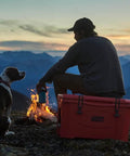 Person and dog sitting by a campfire with a coral cooler in front of a mountain range during sunset.