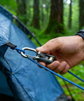 Person securing blue tent with rope and carabiner in rainy forest setting