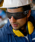 Worker wearing 12 Pack Tinted Safety Glasses ANSI Z87.1 UV400 Eyewear for Men and Women, with hard hat and earmuffs.