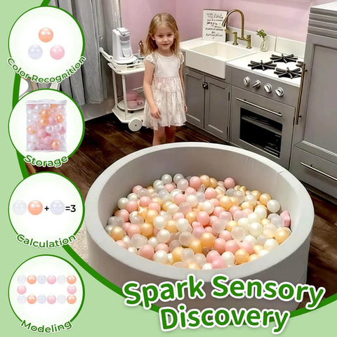 Toddler with gray ball pit filled with plastic balls in pink, gold, pearl white, and clear colors in a kitchen setting.