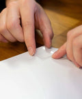 Hands applying Tough And Clear Double Sided Tape Squares For Mounting, 24 Pre-Cut 1-Inch Squares, to a white surface.