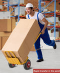 Worker in warehouse using a yellow hand truck with large boxes, wearing blue overalls and white hard hat.
