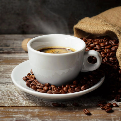 White coffee cup filled with espresso on a wooden table, surrounded by coffee beans and burlap sack in the background.